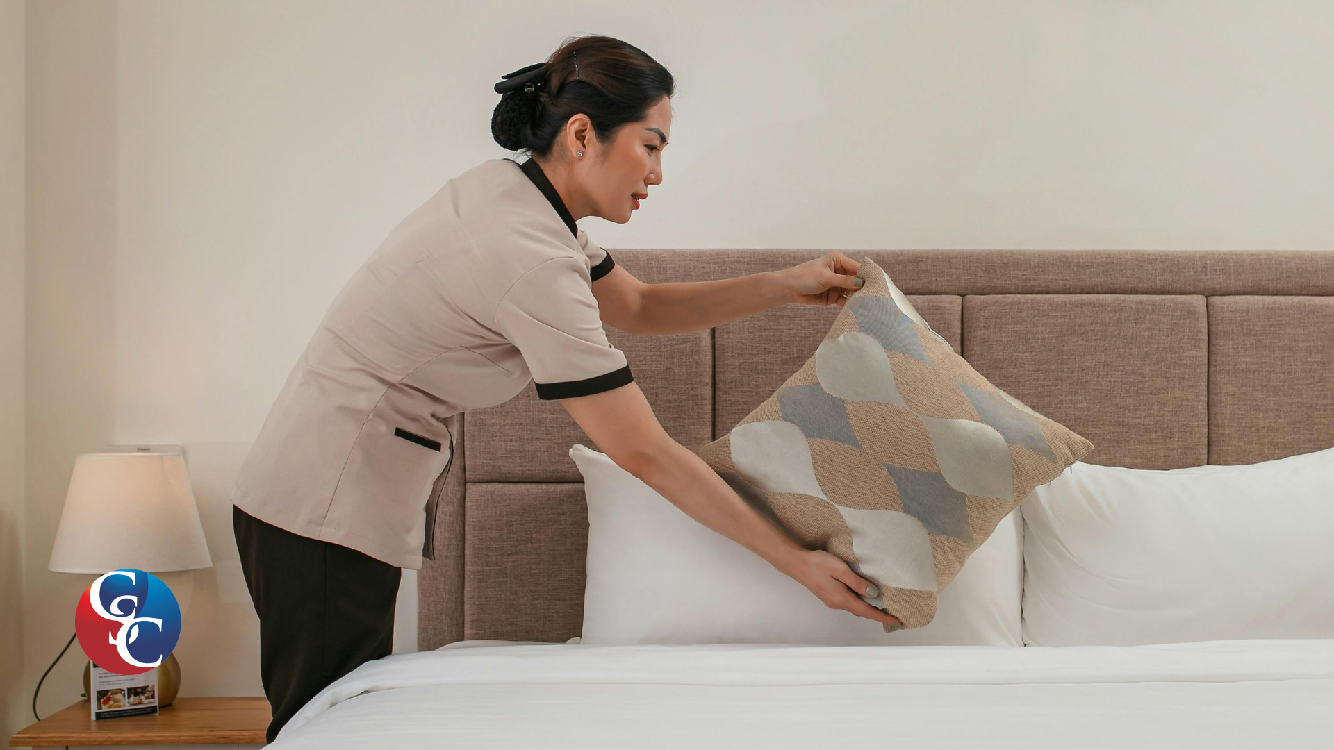A hotel chambermaid in a uniform neatly smoothing down a decorative pillow while making a bed in a modern, well-lit hotel room.