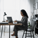 A professional woman in a gray business suit sitting at a modern desk, working on a laptop in a bright, minimalist office space. The image includes a small "GC" logo overlay in the bottom left corner, intended as a cover for a "Payroll Financing 101" blog post.