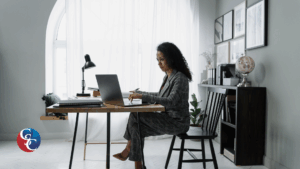 A professional woman in a gray business suit sitting at a modern desk, working on a laptop in a bright, minimalist office space. The image includes a small "GC" logo overlay in the bottom left corner, intended as a cover for a "Payroll Financing 101" blog post.