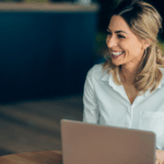 A professional header image for a $100K business loan blog post, featuring a smiling businesswoman in a white shirt working on a laptop in a bright, modern office. The Clear Skies Capital logo is displayed in the bottom left corner.