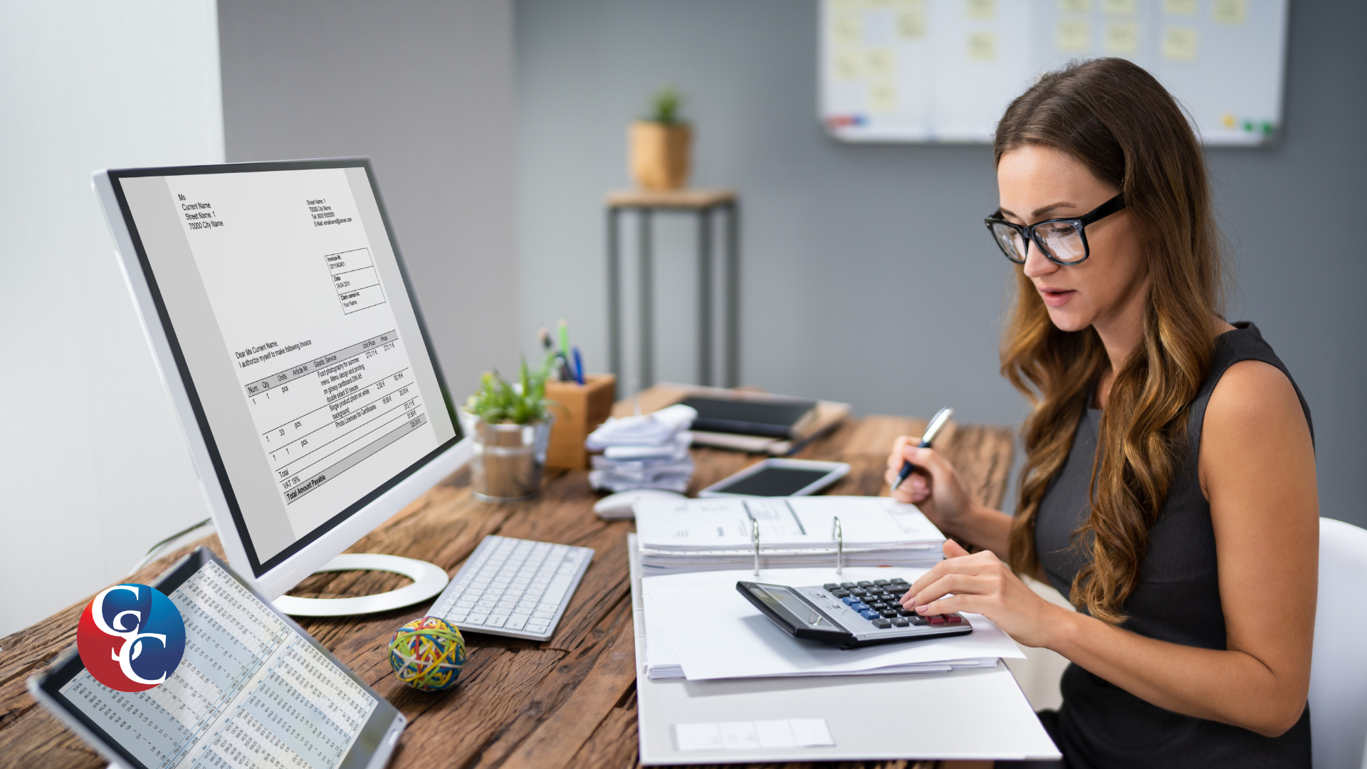 A female accountant with glasses sits at a wooden desk, using a calculator and looking at financial documents in a binder. On her desk is a large desktop monitor displaying financial spreadsheets and data, reflecting a business loan calculation environment.